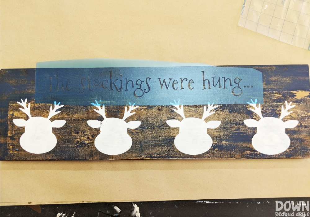 reindeer-stocking-holder-diy-wide-3 Overhead shot of a stencil that says "The stockings were hung..." applied to a wooden board with reindeer faces on it.