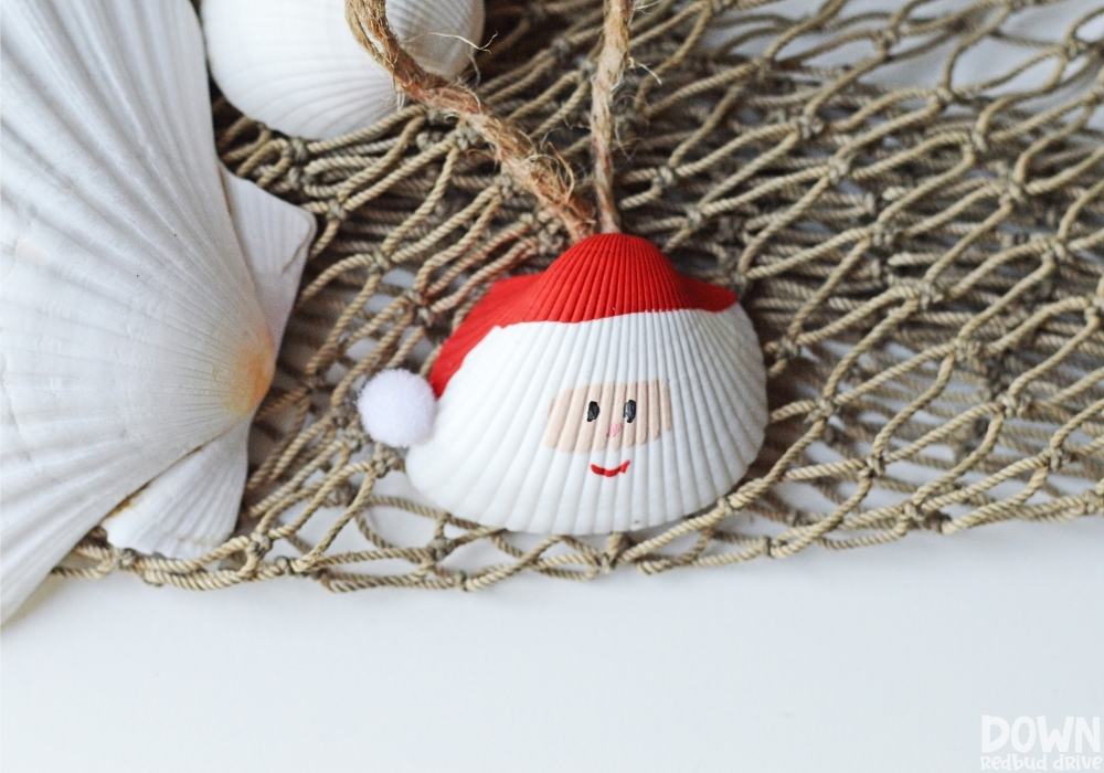santa-seashell-ornament-diy-wide-5 Overhead picture of the finished Santa Seashell Ornament on a fishnet next to unpainted seashells.